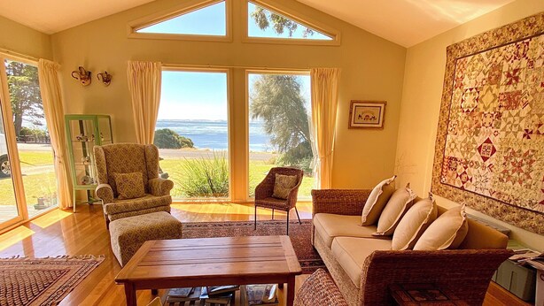 A spacious living room with a high ceiling, large windows, and a view of the outdoors. - Absolute waterfront views, a cosy, country style retreat, set amongst the trees. (Newhaven)