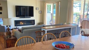 A dining area with a wooden table, chairs, and a bowl of fruit. There is a fireplace and a television in the background. - Absolute waterfront views, a cosy, country style retreat, set amongst the trees. (Newhaven)