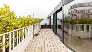 En træterrasse med hvid gelænder, en glasvæg der spejler en bygning, og grøn vegetation på den ene side.