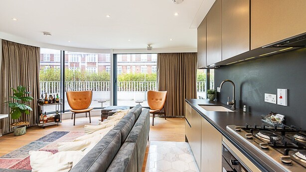 En moderne lejlighed med køkken, to stole, en sofa og en balkon med udsigt til bygninger.