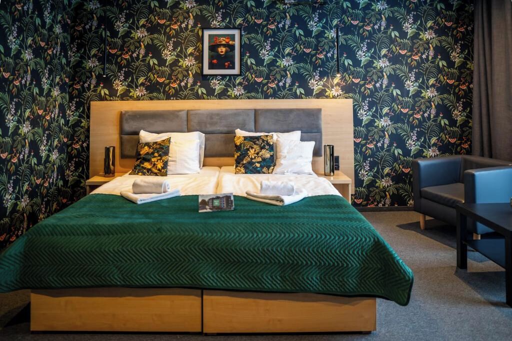 A hotel room with a large bed, a wooden headboard, floral wallpaper, a framed picture on the wall, and a small table with a chair.