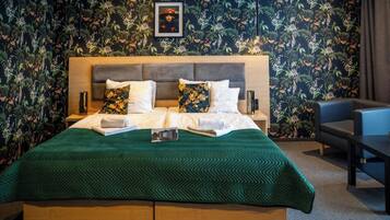 A hotel room with a large bed, a wooden headboard, floral wallpaper, a framed picture on the wall, and a small table with a chair.