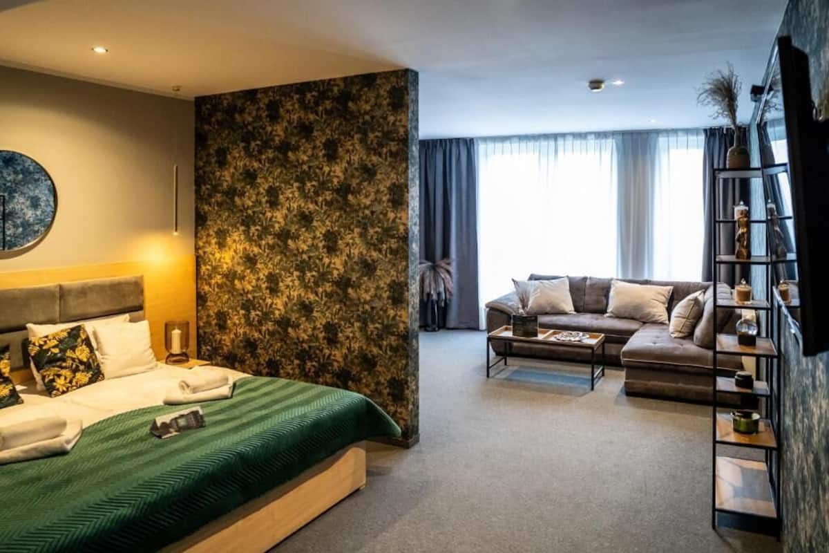 A hotel room with a bed, a sofa, a coffee table, a bookshelf, and a large window with curtains.