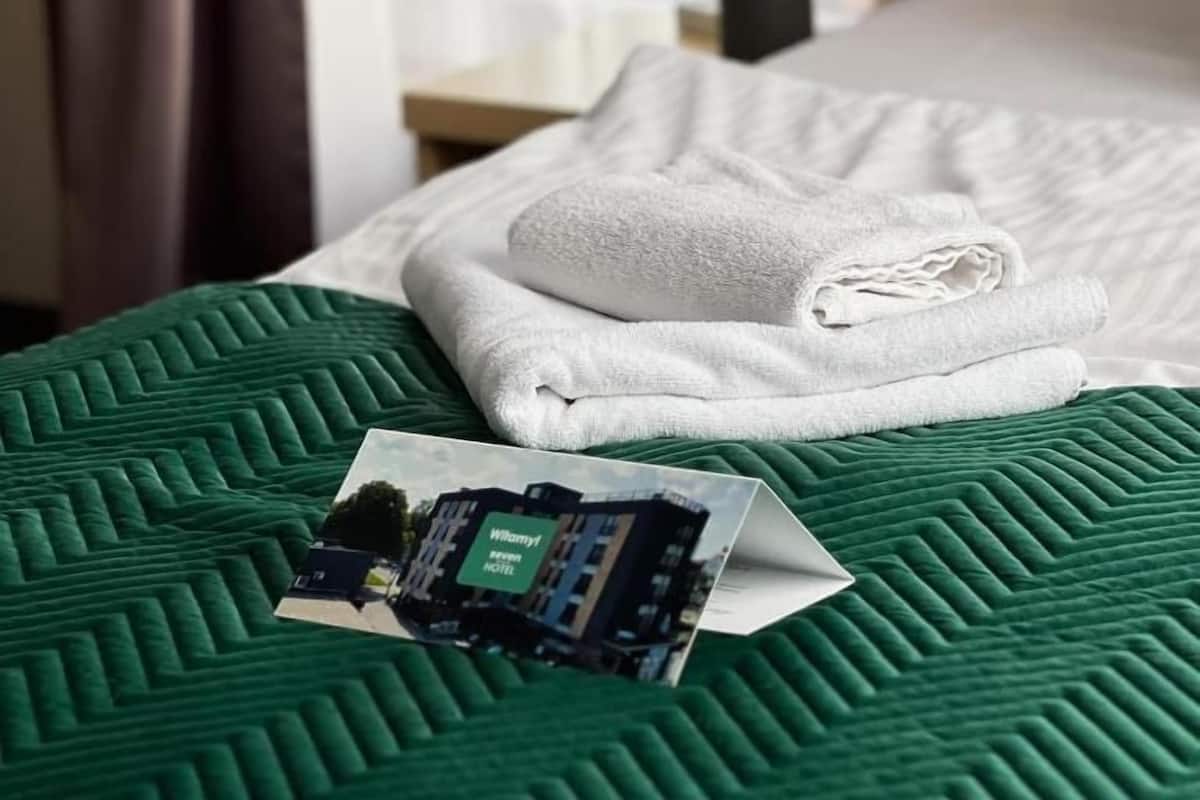 A neatly made bed with a green quilt, a folded white towel, and a printed hotel sign on the bed.