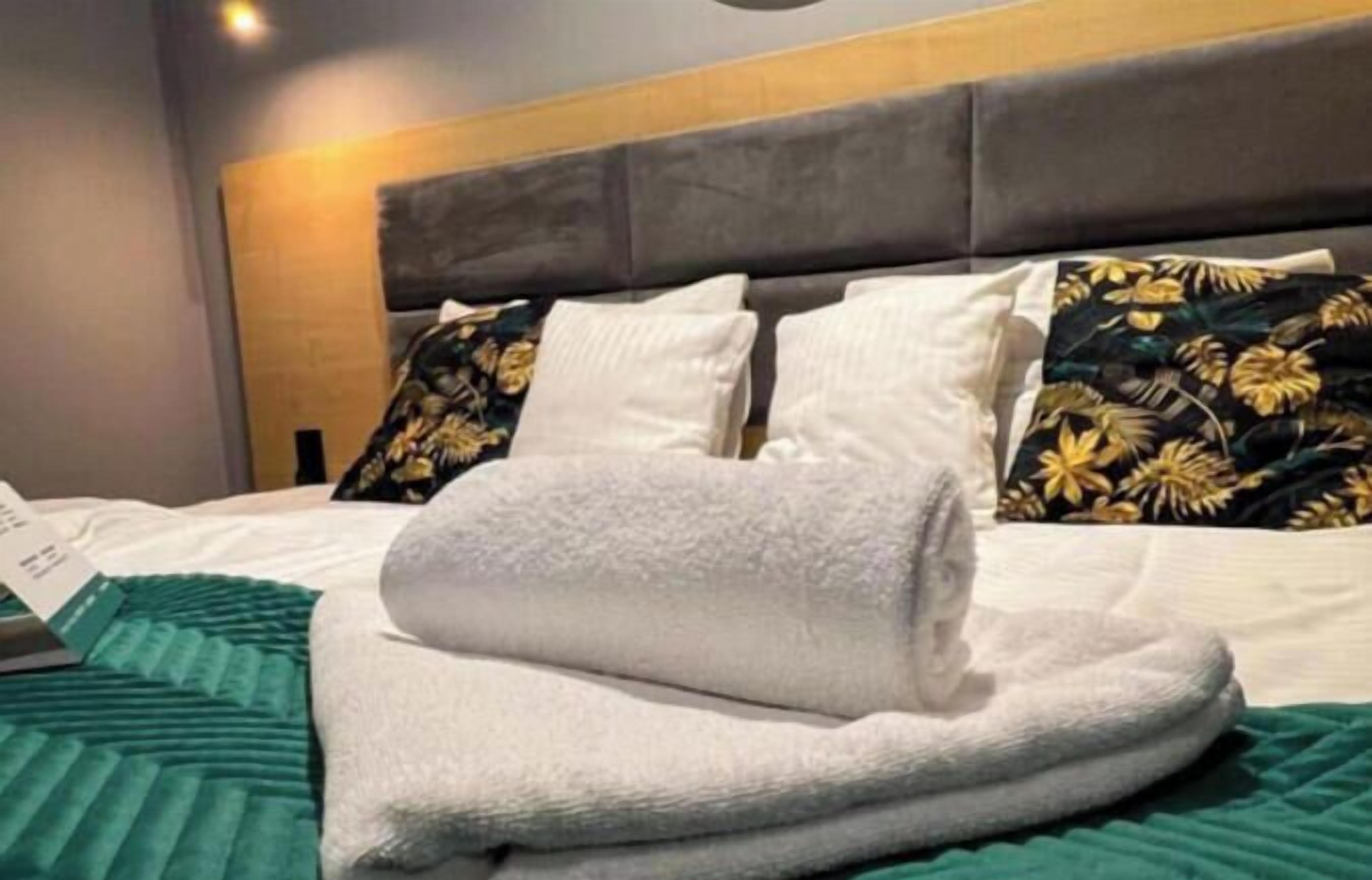 Un lit bien fait, avec des oreillers blancs et une serviette pliée, une couverture verte et un coussin à motifs floraux.