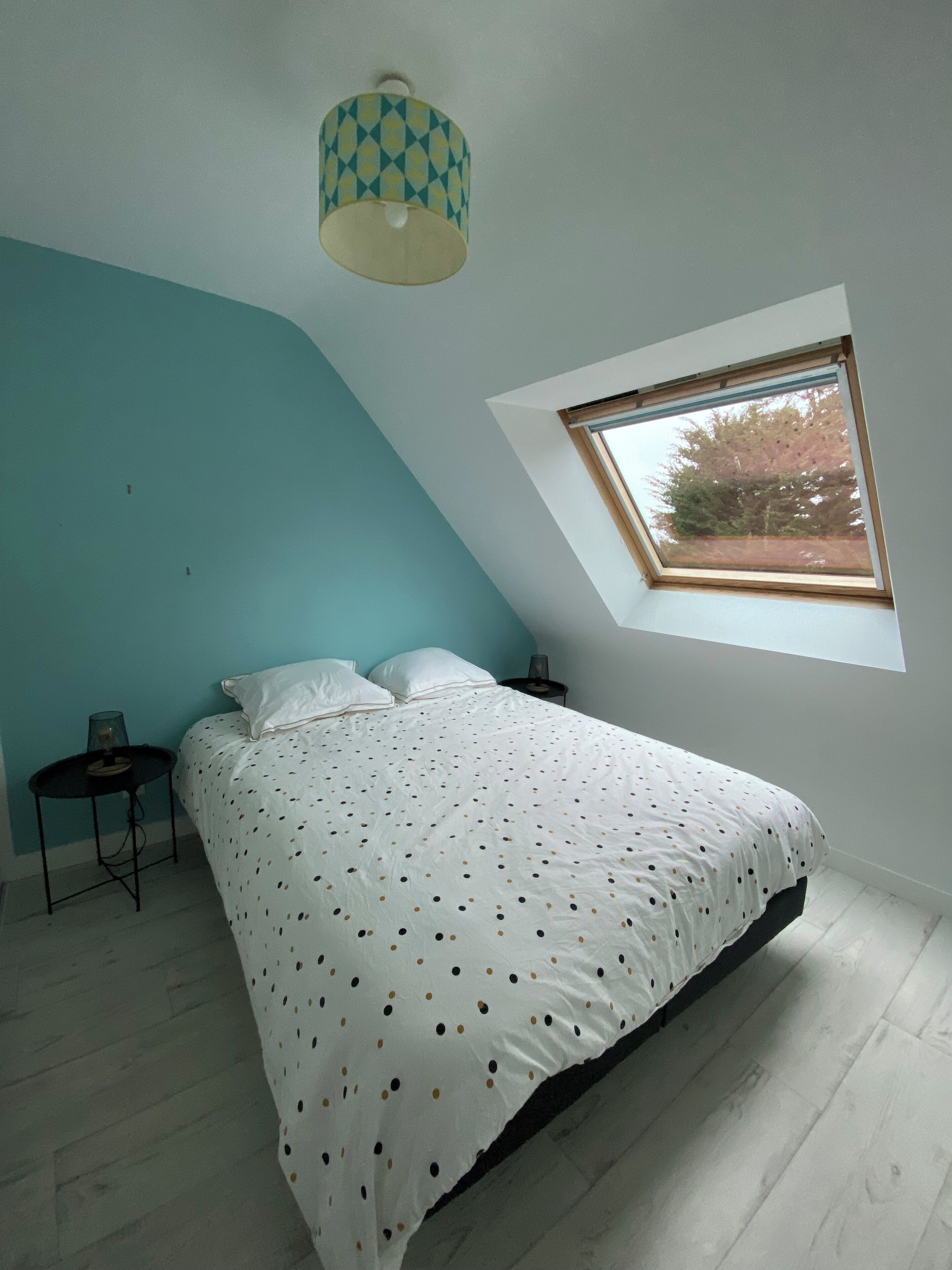 Ein Schlafzimmer mit einem Bett, einem kleinen Tisch, einer Lampe und einem Fenster mit Blick auf Bäume.