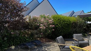 Eine Holzterrasse mit Gartenmöbeln, darunter Stühle und ein Tisch, umgeben von üppiger Bepflanzung und blühenden Pflanzen.