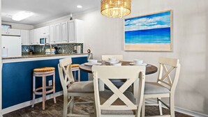Un comedor con una pared azul de acento, sillas blancas, una mesa redonda y un cuadro de una escena de playa.