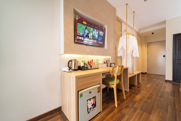 Habitación de hotel con escritorio de madera, refrigerador pequeño, una silla, televisor montado en la pared y una puerta al fondo.