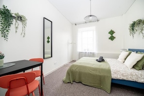 A bedroom with a bed, a desk with chairs, a mirror, and hanging plants.