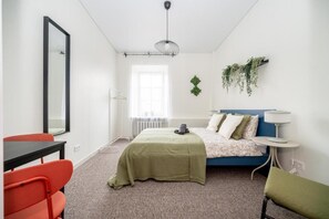 A bedroom with a bed, a desk, chairs, a hanging plant, and a radiator.