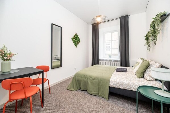 A bedroom with a bed, a desk with chairs, a mirror, and a hanging plant.