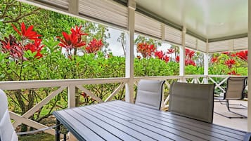 Een overdekt terras met een tafel en stoelen, omgeven door weelderig groen en rode bloemen.