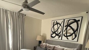 Ein Schlafzimmer mit einem Bett, einem Deckenventilator, einem Nachttisch, einer Lampe und abstrakten Wandbildern.