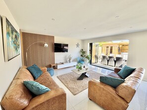 Ein modernes Wohnzimmer mit Ledersofas, einem Flachbildfernseher, einer Wand aus Holzlamellen und Blick auf eine Terrasses.