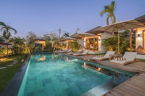 Ein Schwimmbad mit Liegestühlen und Sonnenschirmen, umgeben von Grünflächen und einer Holzterrasse.