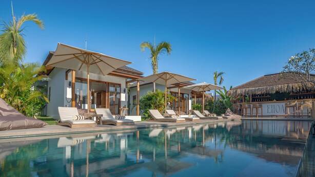 Un área junto a la piscina con sillones de descanso y sombrillas, una estructura con techo de paja y un cielo azul despejado.
