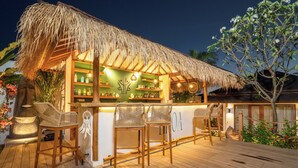 Zona de bar con techo de paja, sillas de madera y una variedad de botellas en estantes.