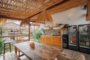 Ein Holzespeisesaal mit Stühlen, einer Pendelleuchte, einer Küchenzeile mit Holzschränken und einem mit Getränken gefüllten Kühlschrank.