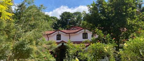 A two-story house with a red-tiled roof, surrounded by lush greenery and trees.