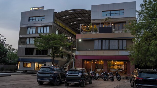 Un edificio de varios pisos con un diseño moderno, que cuenta con un restaurante en la planta baja con terraza, un paseo techado y un estacionamiento con varios vehículos.
