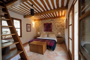 Ein Schlafzimmer mit Steinwand, Holzbalken, einem Bett mit farbenfroher Decke, einer Holzleiter und einem Fenster.