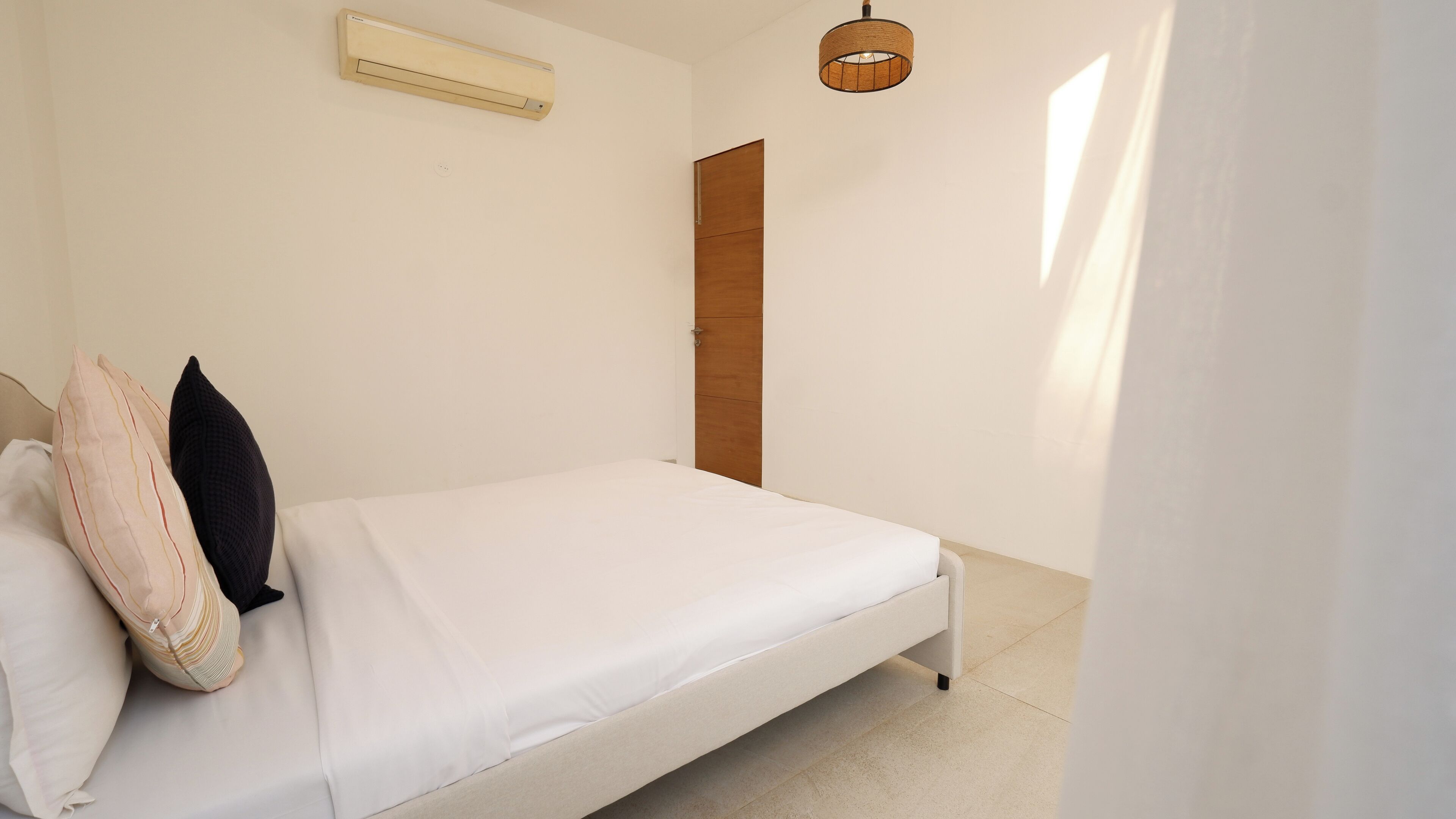 A bedroom with a bed, a wall-mounted air conditioner, a wooden door, and a hanging light fixture.