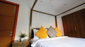 A bedroom with a wooden wardrobe, a bedside table with a plant, and a bed with white bedding and two decorative pillows. - Sun Contemporary by Dia Homes (Nerul)