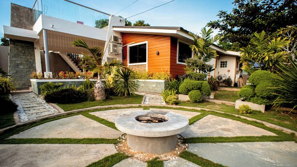 A modern house with a stone fire pit in the center, surrounded by a well-maintained garden with various plants and a stone pathway.