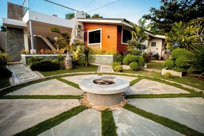 A modern house with a stone fire pit in the center, surrounded by a well-maintained garden with various plants and a stone pathway.