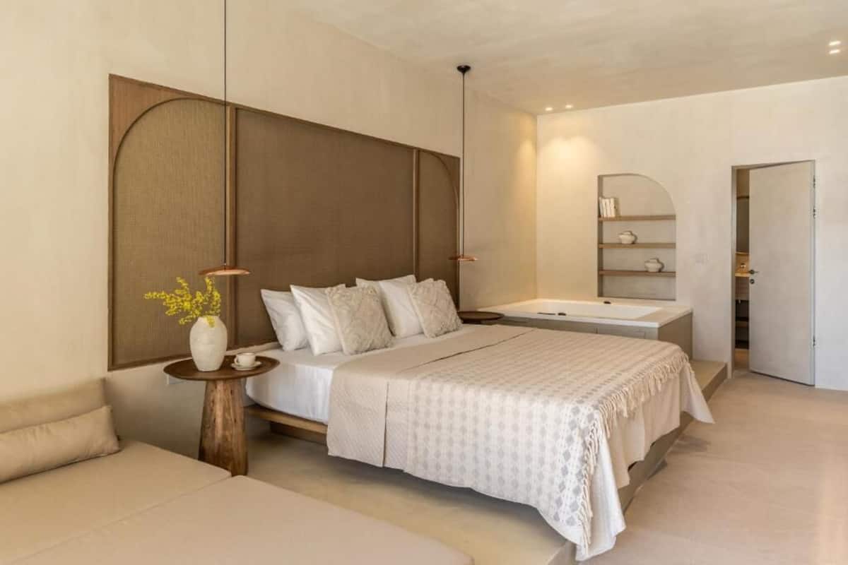 A modern bedroom with a large bed, bedside tables, a small seating area, and a built-in shelf with books and decorative items.