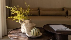 A round wooden table with a white ceramic pitcher and a small bowl containing a melon, set against a beige sofa in a room with a neutral color palette.