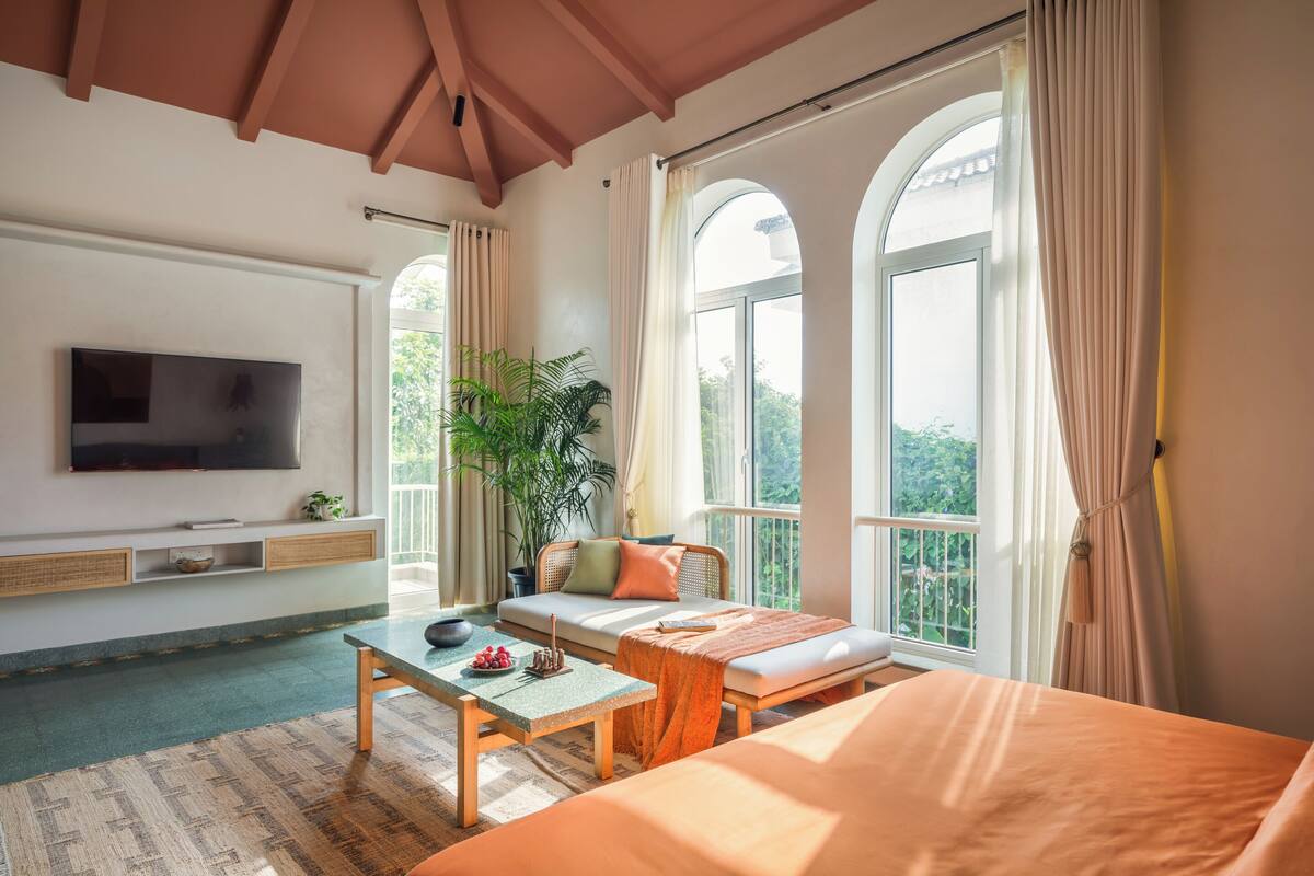 Una habitación con cama, una mesa, televisor y ventanas amplias.