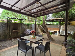 An outdoor patio with a table and chairs, surrounded by greenery and a covered area with a grill.