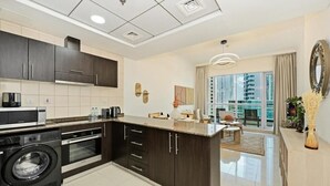 A modern kitchen with dark cabinetry, a built-in washer, and a dining area with a view of high-rise buildings. - Bloomfields Signature 1BR Bay Central We (Dubai)
