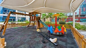 An outdoor playground with a seesaw, a slide, and a climbing structure, surrounded by a wooden fence and palm trees. - Bloomfields Signature 1BR Bay Central We (Dubai)