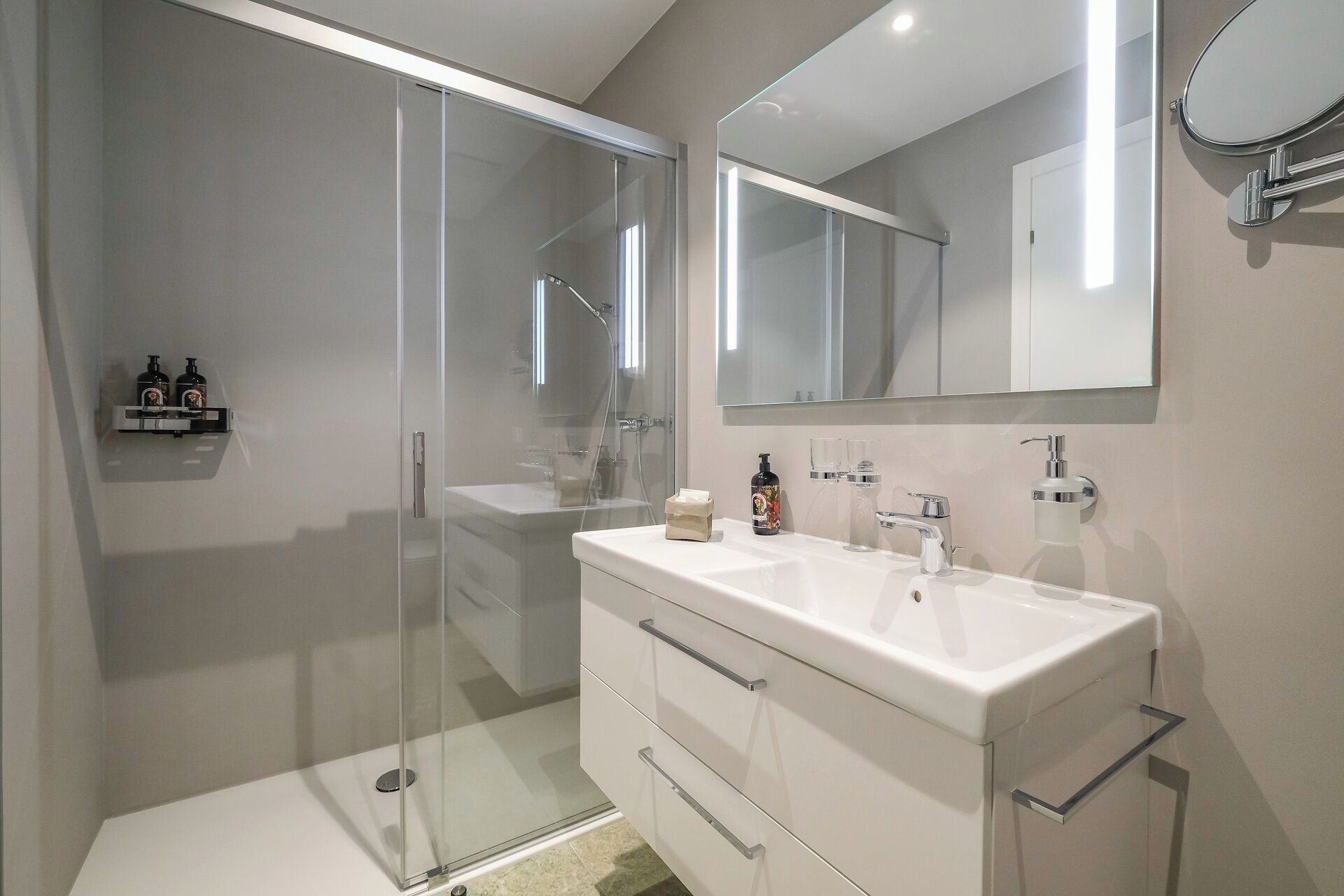 A modern bathroom with a glass-enclosed shower, a white vanity with a large mirror, and a small shelf with toiletries.