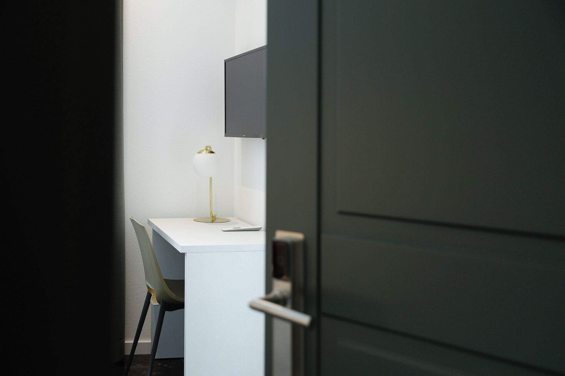 Un espacio de oficina minimalista con un escritorio blanco, una silla, una lámpara de escritorio y una puerta cerrada.