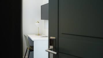 Ein schlicht eingerichteter Arbeitsraum mit weißem Schreibtisch, einem Stuhl, einer Schreibtischlampe und einer geschlossenen Tür.