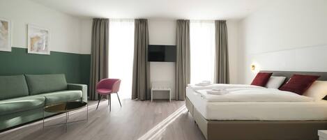 Ein modernes Hotelzimmer mit einem grünen Sofa, einem rosafarbenen Sessel, einem Bett mit weißer Bettwäsche und einem an der Wand befestigten Flachbildfernseher.