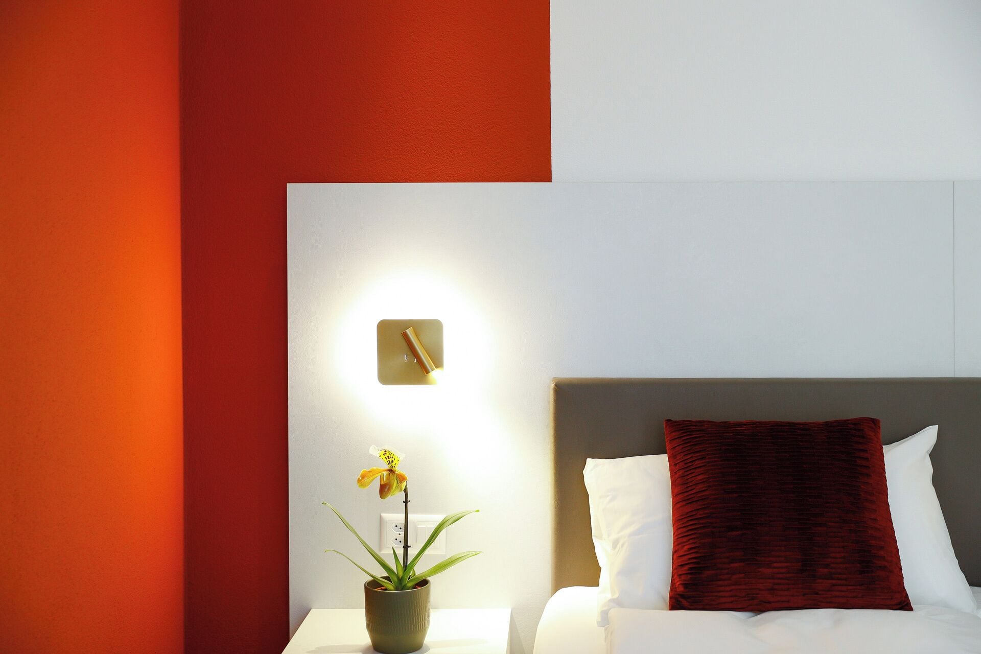 A hotel room with a bed, a nightstand with a potted plant, and a wall-mounted light.