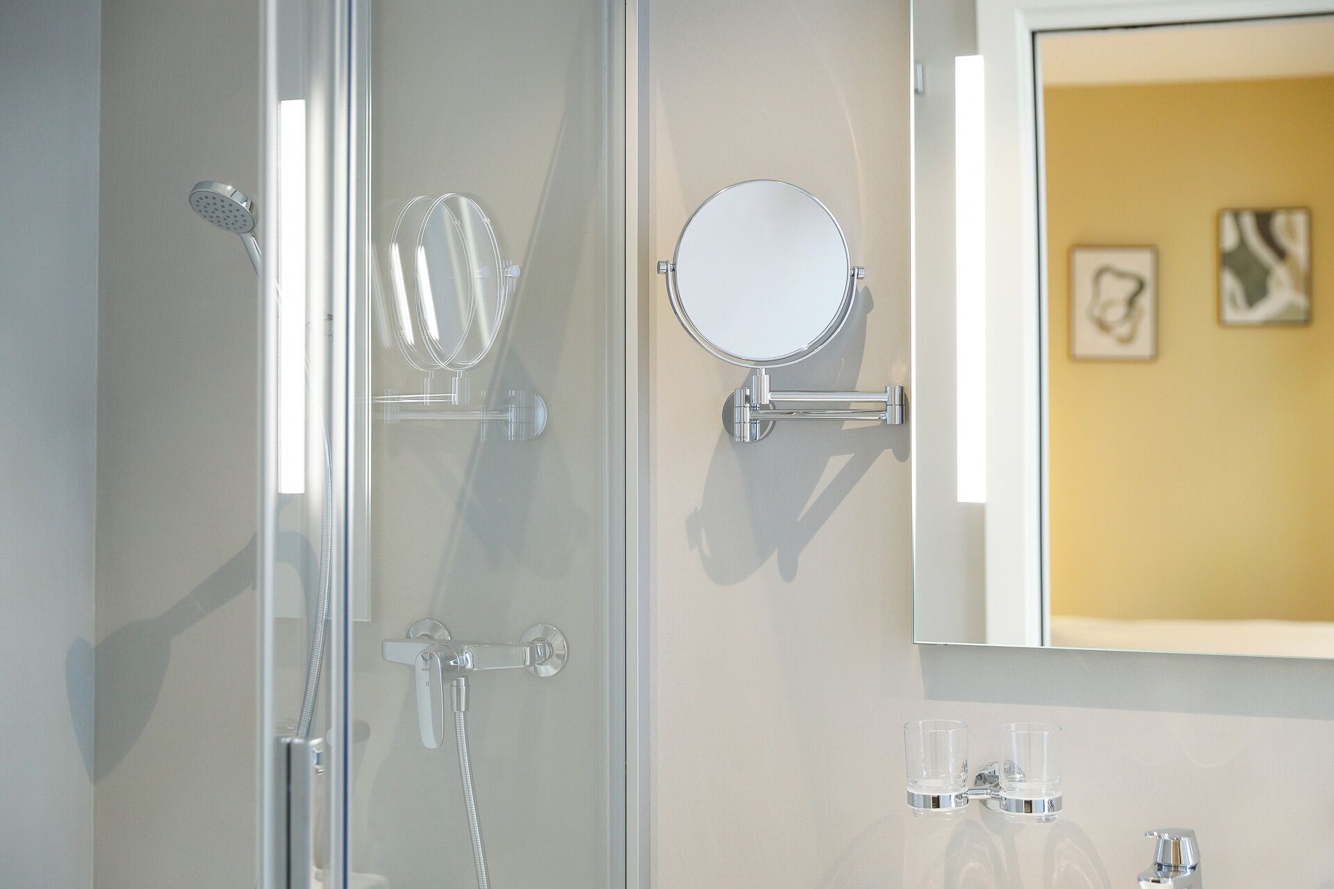 A bathroom with a glass shower enclosure, a handheld showerhead, a wall-mounted mirror, and a sink with two clear glasses.