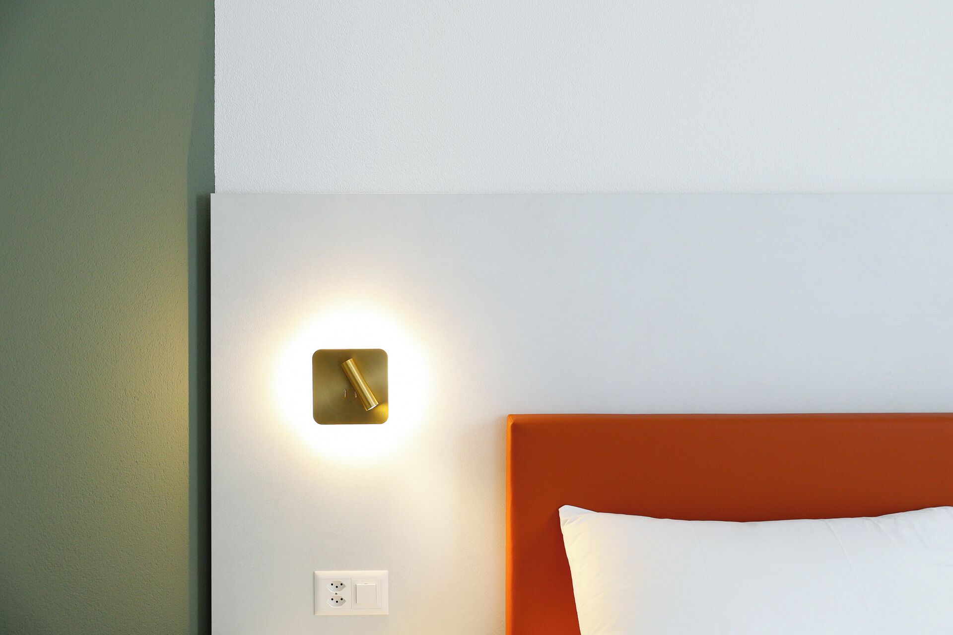 Une chambre d’hôtel avec un mur blanc, une horloge murale de couleur dorée, une tête de lit orange et un oreiller blanc.