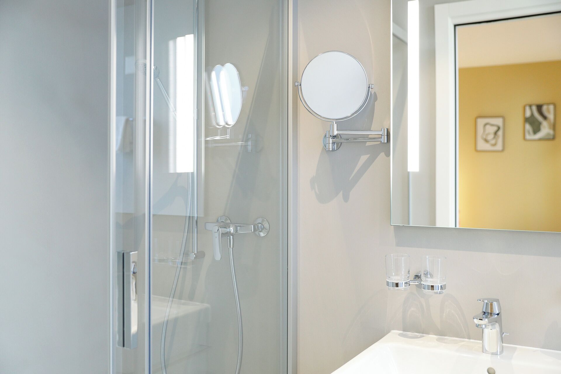 A modern bathroom with a glass shower enclosure, a round mirror, and a white sink with two glass soap dispensers.