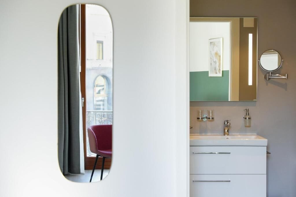 A bathroom with a large mirror, a sink, and a red chair visible through a window.