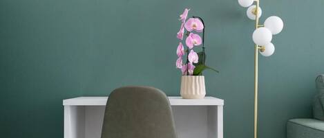 A modern room with a teal accent wall, a white desk, a grey chair, a white vase with pink flowers, and a tall white column with spherical lights.