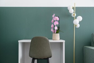 Una habitación moderna con una pared verde azulado, un escritorio blanco, una silla gris, un jarrón blanco con flores rosadas y un alto pilar blanco con luces esféricas.