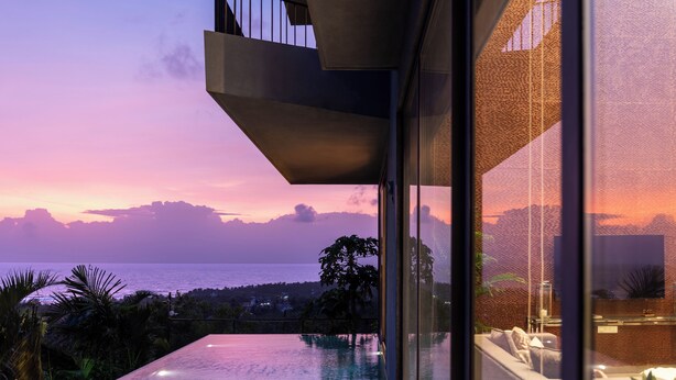 A modern building with a pool, outdoor seating, and a view of the sea at sunset.