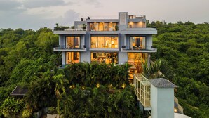 A modern multi-story house with large windows, balconies, and a lush green forest surrounding it.