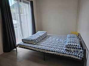 A small, single-bed room with a wooden floor, a window with sheer curtains, and a view of the outside. - Charming 3-bedroom House in Bueng Kum, Thailand (Bangkok)